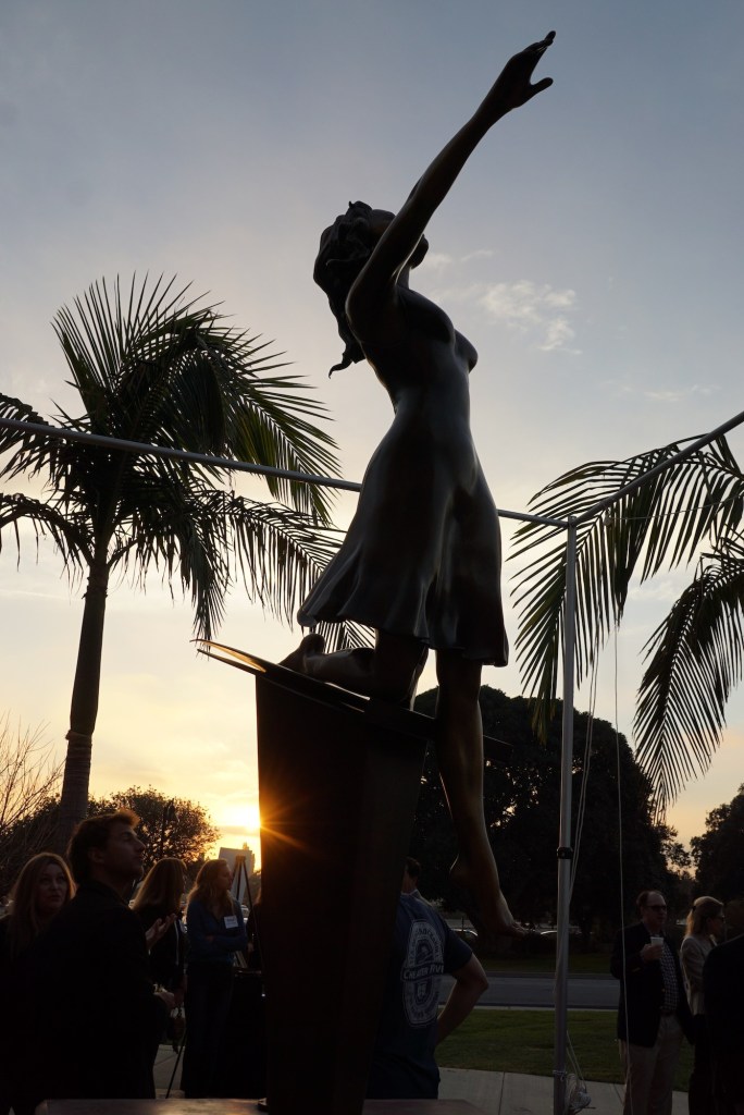The bronze statue of a girl holding a violin with the sun setting behind it.