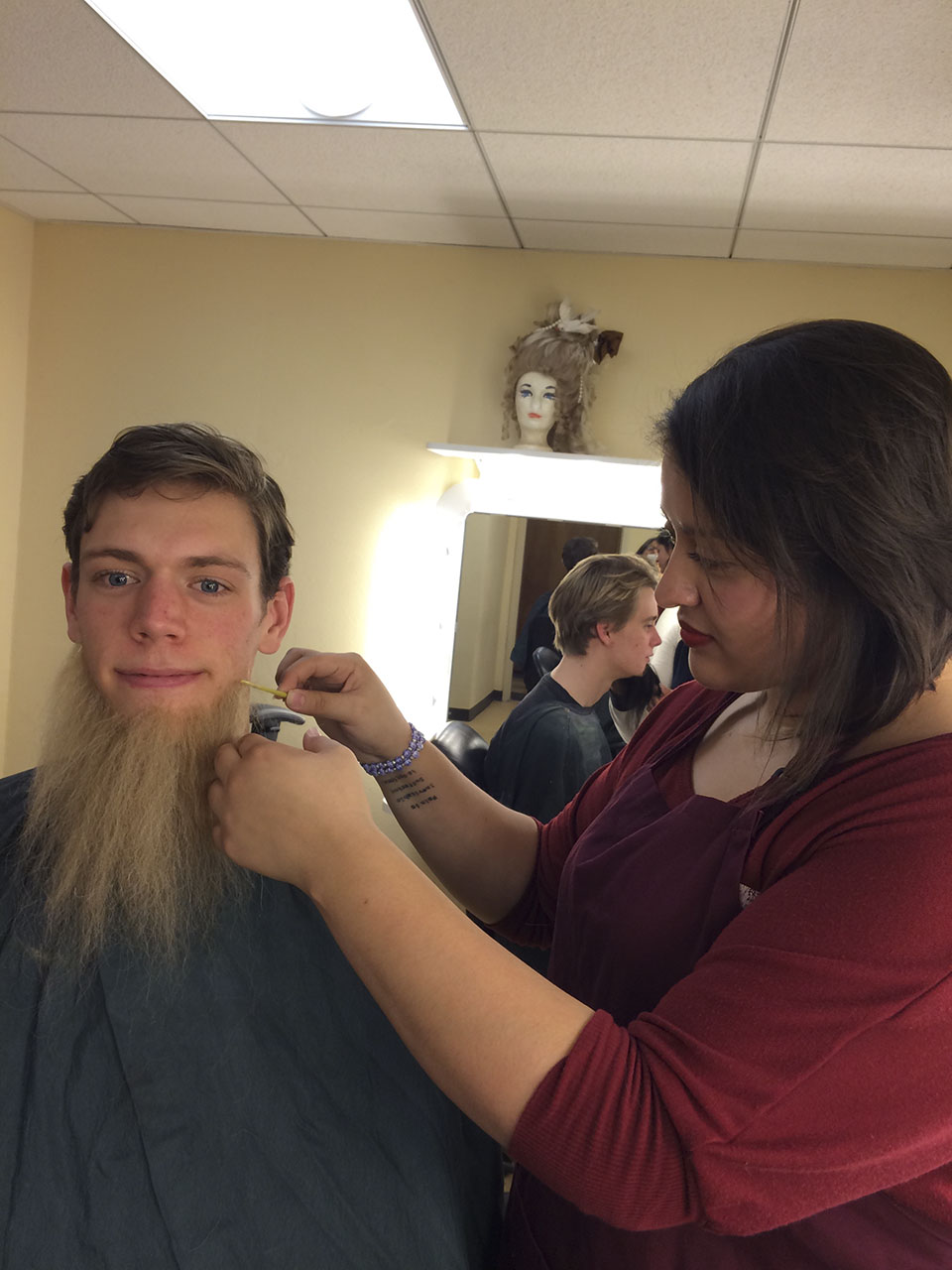 A female student with dark hair applies a false beard to a caucasian male student.