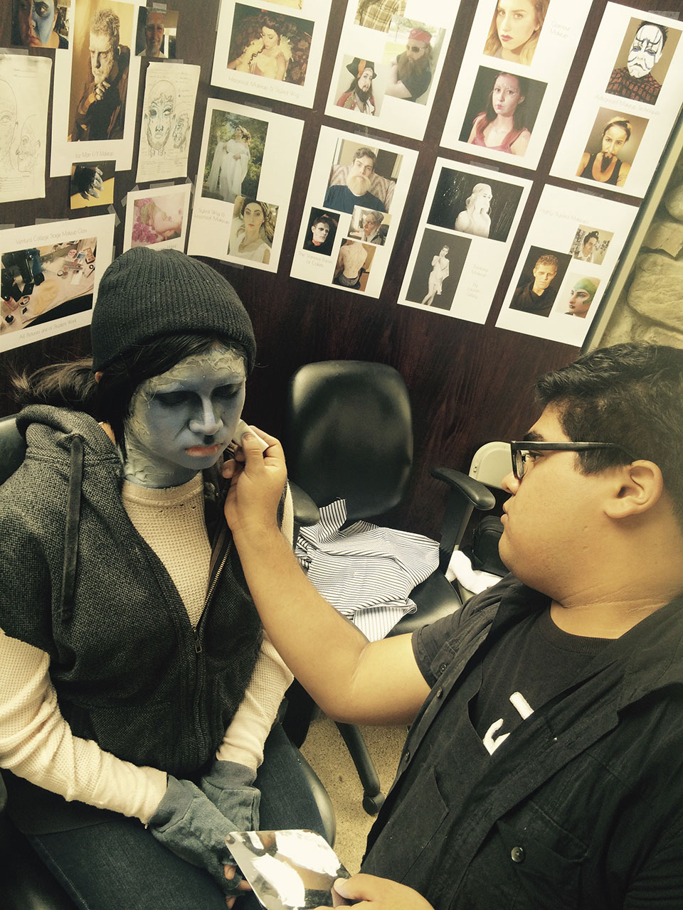 A male student with dark hair applies a monster gray character makeup to a student wearing a black beanie. Makeup images can be seen on the wall behind them.