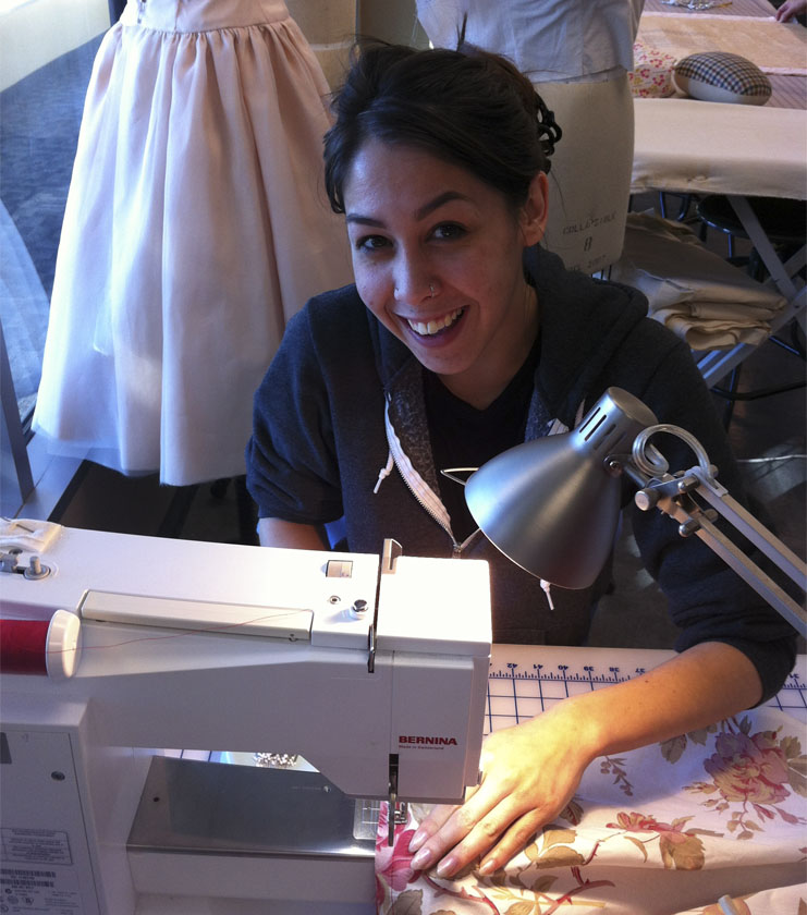 A female student with dark hair sews at a sewing machine and smiles at the camera