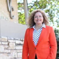Dr. Claudia Lourido-Habib, president of Ventura College, smiles while standing outdoors in front of a stone wall and a “VC” sign. She wears a bright orange blazer, a white patterned blouse, and a pearl necklace. Trees and blue sky are visible in the background.