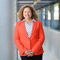 President Lourido-Habib leaning against a column, wearing a VC Orange blazer, black pants, and print blouse
