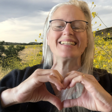 Professor Anna Carlson smiling in font of a field of wildflowers making a heart shape with her hands. 