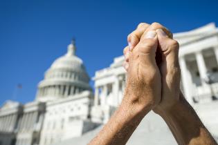 Two hands clasped together in front of a state capital building.
