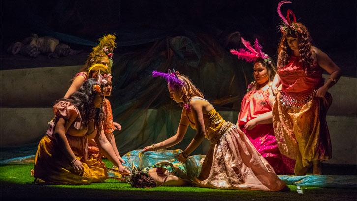 Actors in bright yellow, pink and purple fairy costumes surround an actor on a grassy stage in a Ventura College production of A Midsummer Night's Dream.