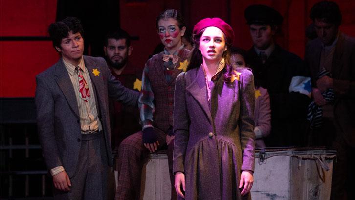 An actress in a red beret and coat is illuminated center stage as actors in suits look on.