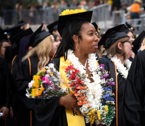 A smiling graduate holds a bouquet and wears colorful leis a